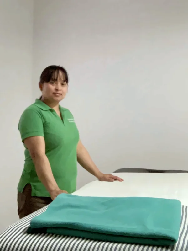 Massør Tin Tin Moe i grøn poloshirt står ved massagebriksen i klinikken, hvor et turkis tæppe ligger klar.